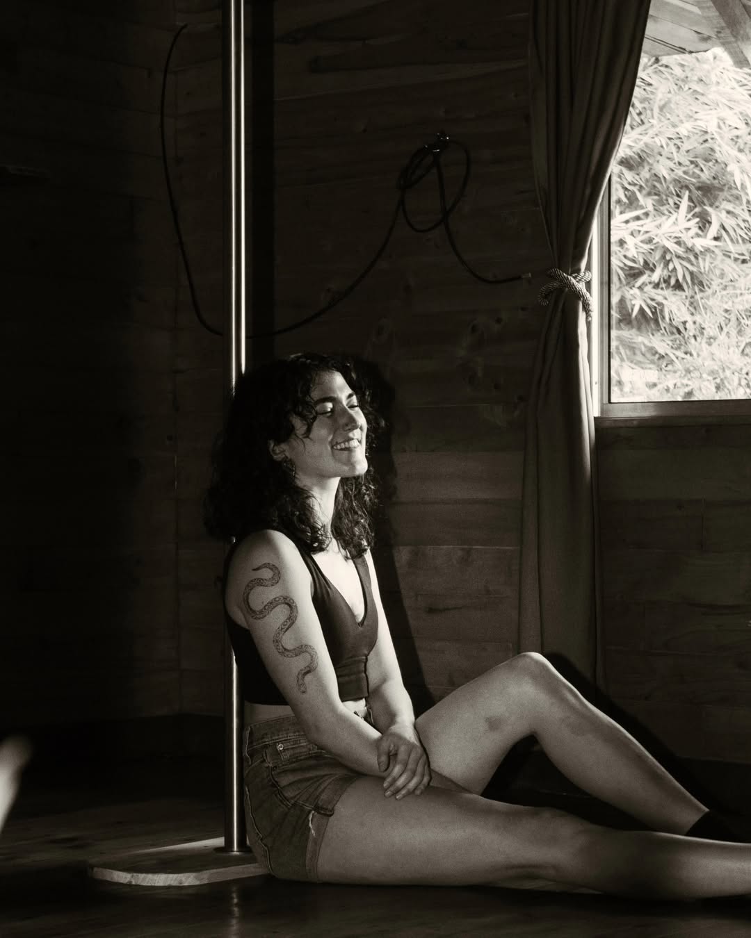 Pilar Waters sitting against a wooden wall in a cabin, sunlight catching her face, snake tattoo visible on her arm.